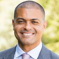 Shereef has shaved hair, smiling in a garden, wearing a gray suit.